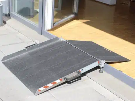 Rollstuhlbrücke mit rutschsicherer Oberfläche, das eine Schwelle überbrückt und einen barrierefreien Zugang ermöglicht. Die Rampe ist zusammenklappbar und hat rot-weiße Markierungen an den Seiten.