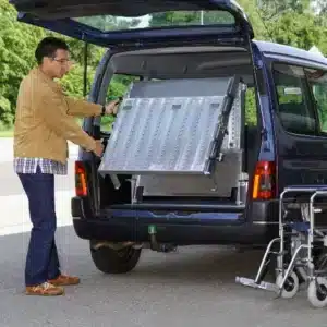 Mann mit brauner Jacke, der eine Rampe aus dem Kofferraum eines blauen Fahrzeugs ausklappt. Die Rampe ist aus Metall und dient zur Unterstützung von Mobilitätshilfen.