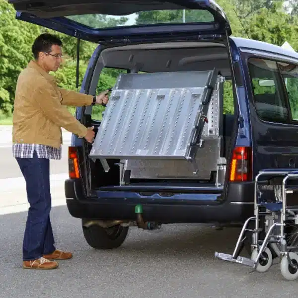 Mann mit brauner Jacke, der eine Rampe aus dem Kofferraum eines blauen Fahrzeugs ausklappt. Die Rampe ist aus Metall und dient zur Unterstützung von Mobilitätshilfen.