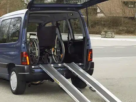 Ein blauer Vans mit offenen Heckklappen, der eine Rampe zeigt, die für den Zugang eines Rollstuhls ins Fahrzeug dient. Im Inneren steht ein Rollstuhl auf der Rampe.