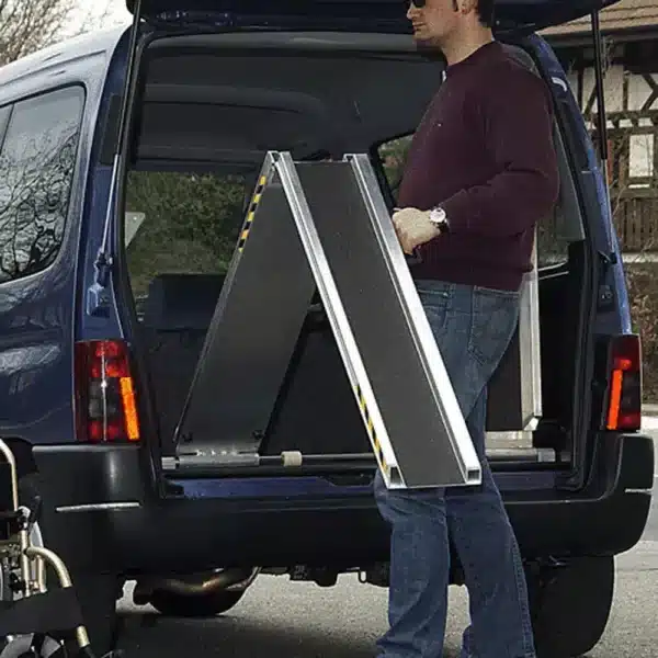 Mann lädt eine Klapprampe in den Kofferraum eines Busses, neben einem Rollstuhl. Kleidung: Dunkelblauer Pullover, Jeans.