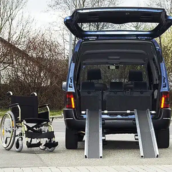 Ein blauer Van mit geöffnetem Heck, in dem eine Rampe zum Einladen eines Rollstuhls angebracht ist. Ein Rollstuhl steht neben dem Fahrzeug auf einer gepflasterten Fläche.