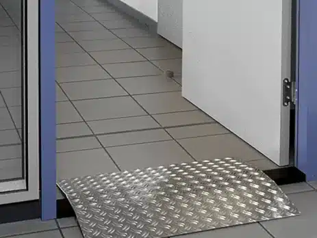 Eine Aluminium-Schwellenbrücke für barrierefreien Zugang, platziert vor einer Tür auf einem Fliesenboden.