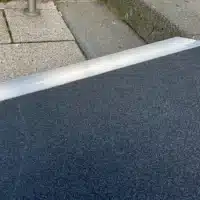 Nahaufnahme einer Rampe aus grauem Material, die auf einen gepflasterten Boden trifft. Die Rampe hat eine leicht erhöhte Kante und ist für barrierefreies Transports geeignet.