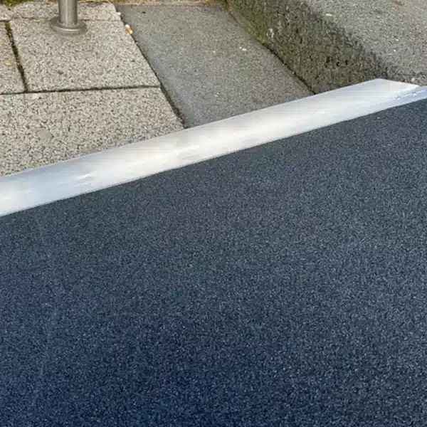 Nahaufnahme einer Rampe aus grauem Material, die auf einen gepflasterten Boden trifft. Die Rampe hat eine leicht erhöhte Kante und ist für barrierefreies Transports geeignet.