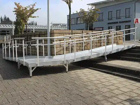 Rollstuhlgerechte Rampe über eine Ecke mit Metallgeländer, die zu einem Eingangsbereich führt, umgeben von mehreren Bäumen und einer Mauer aus Steinen.
