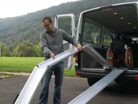 Mann positioniert eine Rampe zum Hineinfahren eines Rollstuhls in einen Van. Im Hintergrund eine grüne Landschaft.