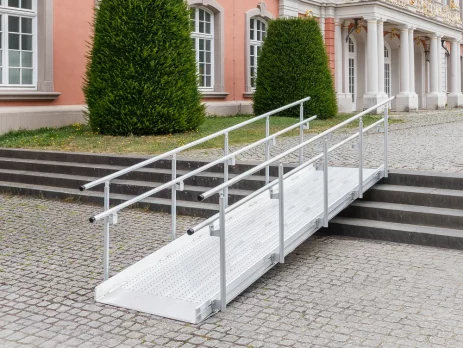 Eine Rollstuhlrampe aus Metall mit Geländer ist über einer Treppe vor einem historischen Gebäude mit großen Fenstern installiert.