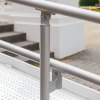 Nahaufnahme eines Metallhandlaufs und eines Stützpfostens an einer für Rollstuhlfahrer zugänglichen Rampe im Freien in der Nähe von Stufen und einem Gehweg.