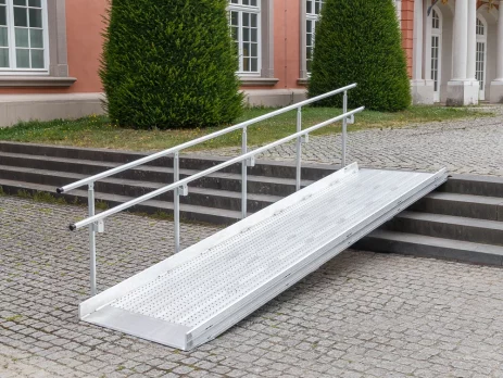 Vor einem historischen Gebäude mit großen Fenstern und Sträuchern ist eine Rollstuhlrampe aus Metall mit Handläufen über Steinstufen angebracht.