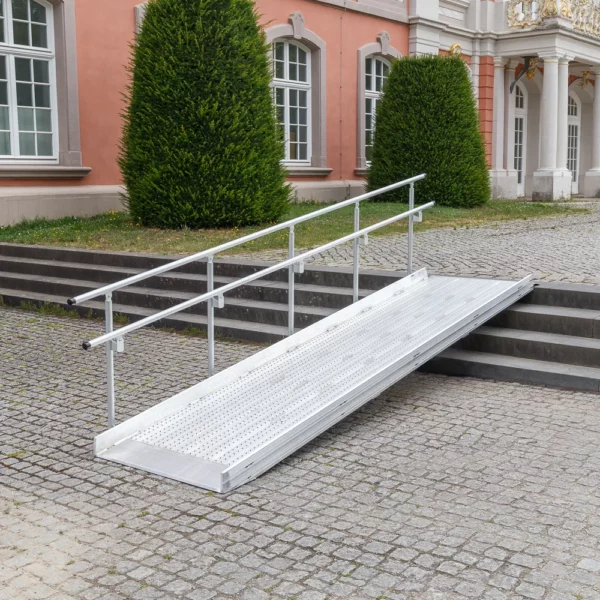 Vor einem historischen Gebäude mit großen Fenstern und Sträuchern ist eine Rollstuhlrampe aus Metall mit Handläufen über Steinstufen angebracht.
