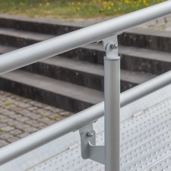 Nahaufnahme eines Metallgeländers neben einer Rollstuhlrampe und einer Treppe im Freien mit Rasen im Hintergrund.