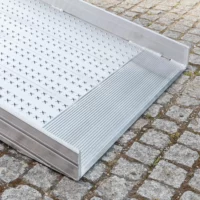Eine Rollstuhlrampe aus Metall mit einer strukturierten Oberfläche ist mit der unteren Auflage auf einem Kopfsteinpflaster platziert.