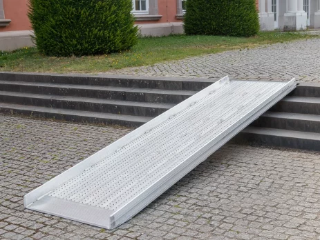 Vor einem Gebäude mit großen Fenstern und gestutzten Hecken ist eine Rollstuhlrampe über Steinstufen angebracht.