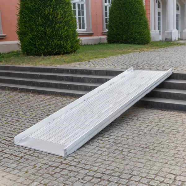 Vor einem Gebäude mit großen Fenstern und gestutzten Hecken ist eine Rollstuhlrampe über Steinstufen angebracht.