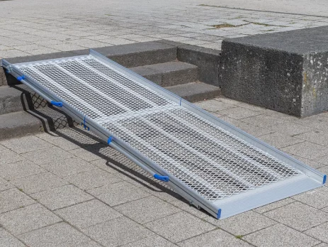 Eine tragbare Rollstuhlrampe aus Aluminium ist über drei Stufen auf einem gepflasterten Außengelände angebracht.