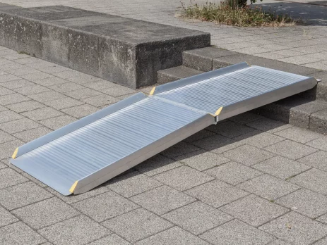 Eine klappbare Rollstuhlrampe liegt auf Stufen und auf einem gepflasterten Gehweg im Freien und ermöglicht Rollstuhlfahrern den Zugang.