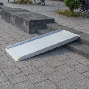 Eine tragbare Aluminium-Rollstuhlrampe liegt auf einer Treppe mit 2 Stufen um den barrierefreien Zugang vom gepflasterten Platz auf den höher liegenden Gehweg zu ermöglichen.