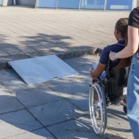 Eine Person im Rollstuhl wird von einer Hilfsperson zu einer tragbaren Rollstuhlrampe aus Aluminium geschoben