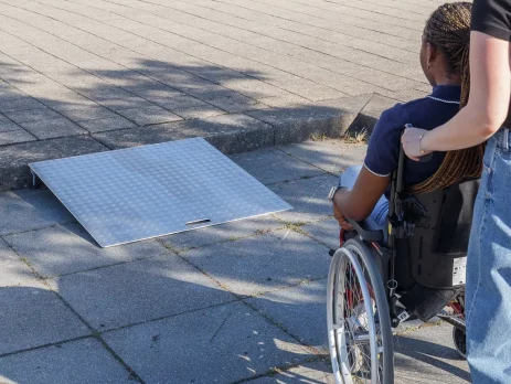 Eine Person im Rollstuhl wird von einer Hilfsperson zu einer tragbaren Rollstuhlrampe aus Aluminium geschoben
