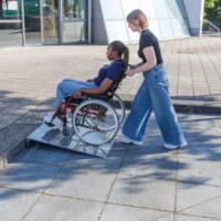 Eine Frau schiebt eine andere Frau im Rollstuhl über eine Rollstuhlrampe aus Aluminium auf den Bürgersteig vor einem modernen Gebäude.