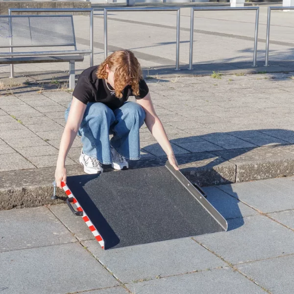 Eine Person richtet eine tragbare Aluminium-Rollstuhlrampe vor einer einzelnen Stufe im Freien auf einer gepflasterten Fläche aus.