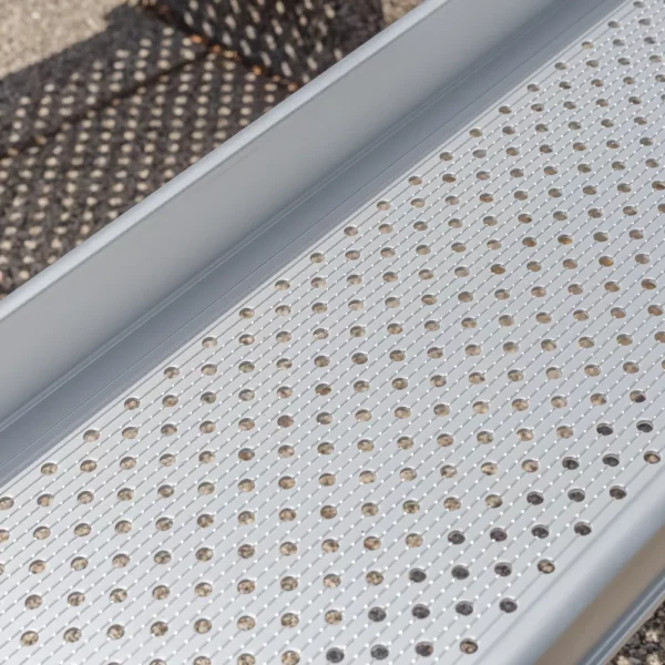 Nahaufnahme der Fahrfläche einer Rollstuhlrampenschiene mit gelochter und längsgeriffelter Oberfläche mit Unterbrechung der Riffel. Die Rampe ist aus Aluminium.