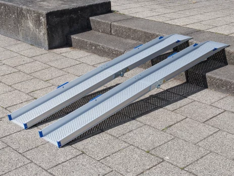 Zwei tragbare Rollstuhlrampen Schienen aus Aluminium sind über eine Außentreppe aus 2 Stufen auf einen gepflasterten Platz gelegt.
