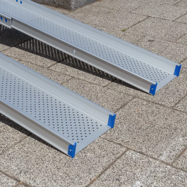Nahaufnahme von zwei unteren Enden der Schienen einer Rollstuhlrampen aus Aluminium liegen an einem sonnigen Tag im Freien auf einer gepflasterten Fläche.
