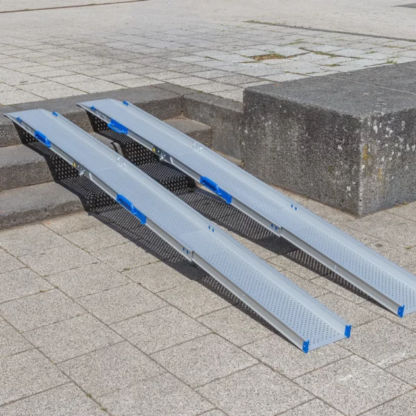 Zwei tragbare Rollstuhlrampen Schienen liegen auf einer Außentreppe und ermöglichen den Zugang zwischen verschiedenen Ebenen einer gepflasterten Fläche.