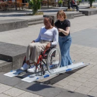 Eine Frau schiebt eine andere Frau im Rollstuhl eine tragbare Rollstuhlrampe auf einem städtischen Bürgersteig hinauf.