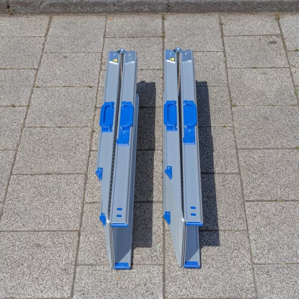 Zwei zusammen geklappte und zusammen geschobene Rollstuhlrampen Schienen aus Aluminium mit blauen Griffen liegen nebeneinander auf einem Gehweg.