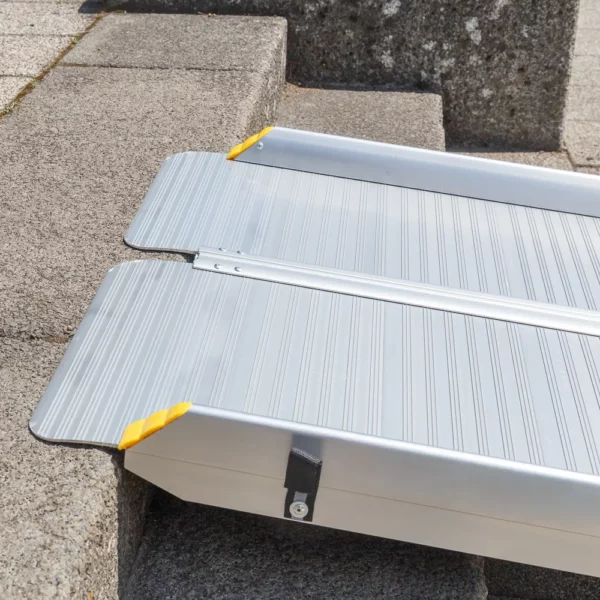 Nahaufnahme des oberen Auflagewinkels einer tragbaren Rollstuhlrampe aufliegend auf der Kante einer Betonstufe mit Darstellung der rutschsicher geriffelten Fahrfläche und des Klappscharniers.