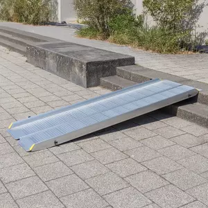 Eine tragbare Rollstuhlrampe aus Aluminium überbrückt eine kurze Außentreppe von 2 Stufen auf einem gepflasterten Gehweg.