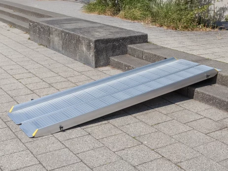 Eine tragbare Rollstuhlrampe aus Aluminium überbrückt eine kurze Außentreppe von 2 Stufen auf einem gepflasterten Gehweg.