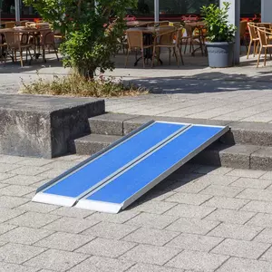 Eine tragbare blaue Rollstuhlrampe wird über Stufen liegt und den Zugang für Rollstuhlfahrer zu einem Sitzbereich im Freien ermöglicht.