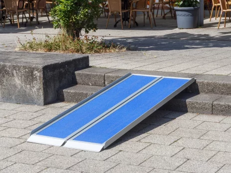 Eine tragbare blaue Rollstuhlrampe wird über Stufen liegt und den Zugang für Rollstuhlfahrer zu einem Sitzbereich im Freien ermöglicht.