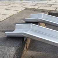 Detailaufnahme der Auflageflächen von zwei Schienen einer Rollstuhlrampe aus Aluminium mit geriffelter Fahrfläche auf der obersten Stufe einer Treppe im Freien.
