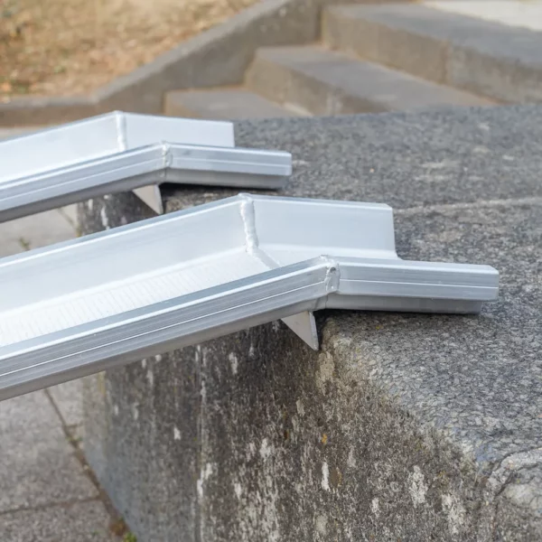 Seitliche Detailaufnahme der Auflageflächen von zwei Schienen einer Rollstuhlrampe aus Aluminium auf der obersten Stufe einer Treppe im Freien.