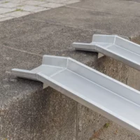 Seitliche Detailaufnahme der Auflageflächen von zwei Schienen einer Rollstuhlrampe aus Aluminium auf der obersten Stufe einer Treppe im Freien.