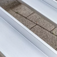 Nahaufnahme von zwei Schienen einer Aluminium-Rollstuhlrampe mit geriffelter Fahrfläche über einer Treppe im Außenbereich.