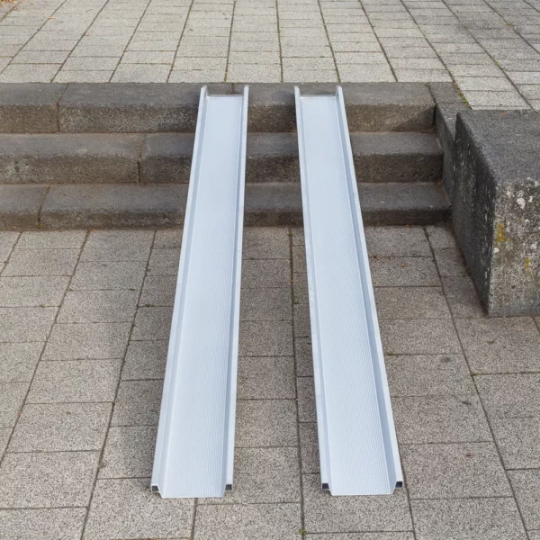 Zwei parallel platzierte tragbare Aluminiumschienen einer Rollstuhlrampe über drei Treppenstufen im Außenbereich vom unteren Ende aus fotografiert.