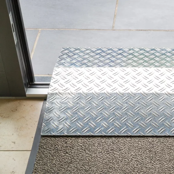 Eine Rollstuhlrampe aus Aluminium mit geriffelter Oberfläche führt von einer Fußbodenmatte zu einem gefliesten Außenbereich.