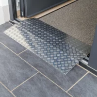 Eine Schwellenrampe aus Aluminium überbrückt eine Türkante und einem gefliesten Boden und einem mit Fußmstte ausgelegten Raum innerhalb einer Türöffnung.