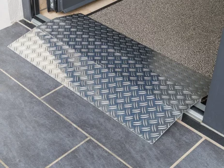Eine Schwellenrampe aus Aluminium überbrückt eine Türkante und einem gefliesten Boden und einem mit Fußmstte ausgelegten Raum innerhalb einer Türöffnung.