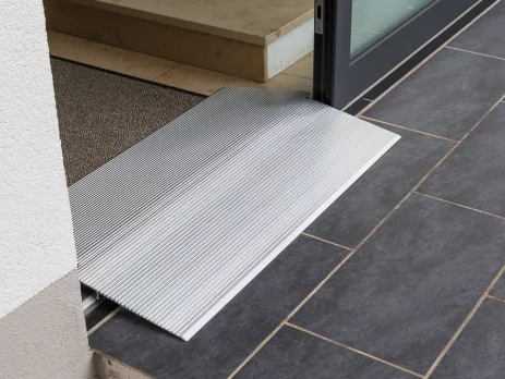 Eine hohe Schwellenbrücke aus Aluminium überbrückt eine Türschwelle zwischen einer Innentreppe und einer gefliesten Außenfläche.