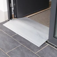 Eine Schwellenbrücke aus Aluminium  liegt an der Schwelle einer Türöffnung über dunklen Bodenfliesen neben einer Fußmatte.