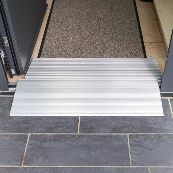 Zwei Schwellenbrücken aus Alumimium  für Rollstühle an einem Türeingang mit dunklem Fliesenboden und einer strukturierten Matte im Inneren.