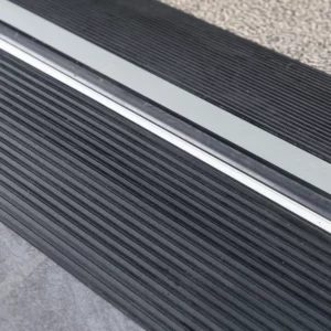 Nahaufnahme der Oberfläche zweier schwarzer Schwellenkeile mit Rillen neben einer Türkante und einem Teppichboden.