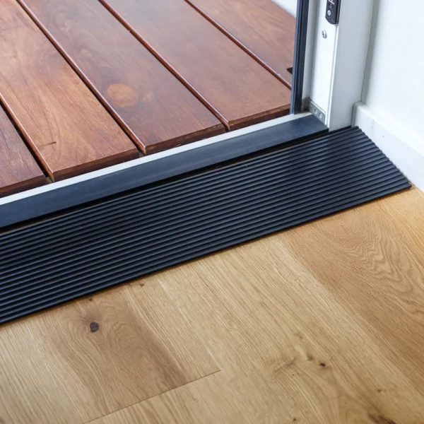 Eine Rampe aus schwarzem Gummi verbindet einen hölzernen Innenboden durch eine offene Tür mit einer Außenterrasse.